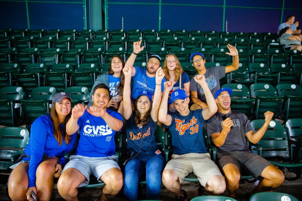 Group photo of people wearing GVSU and Tigers outfits cheering in stands at Comerica Park